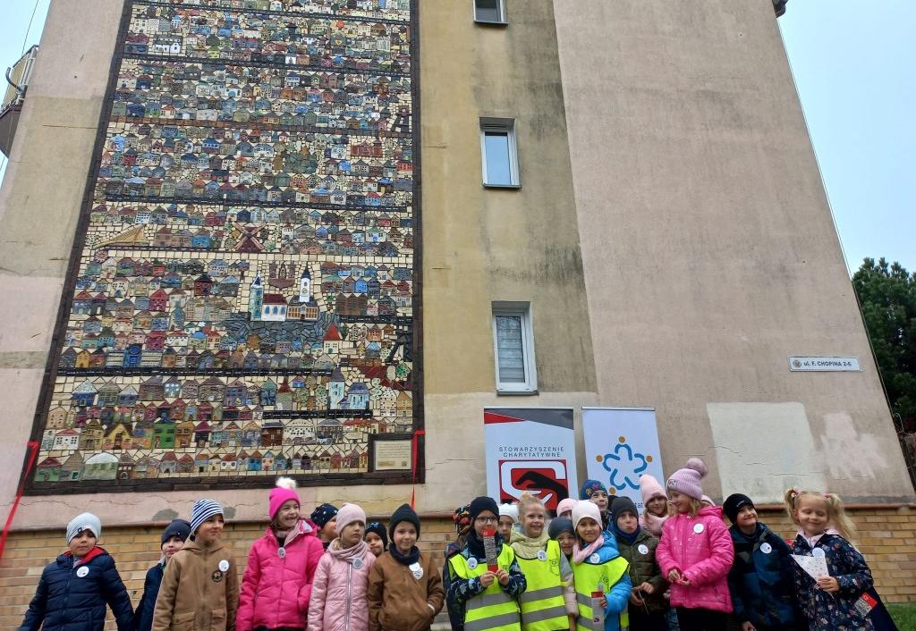 Grupa dzieci stoi na tle ceramicznej mozaiki przymocowanej na ścianie bloku.