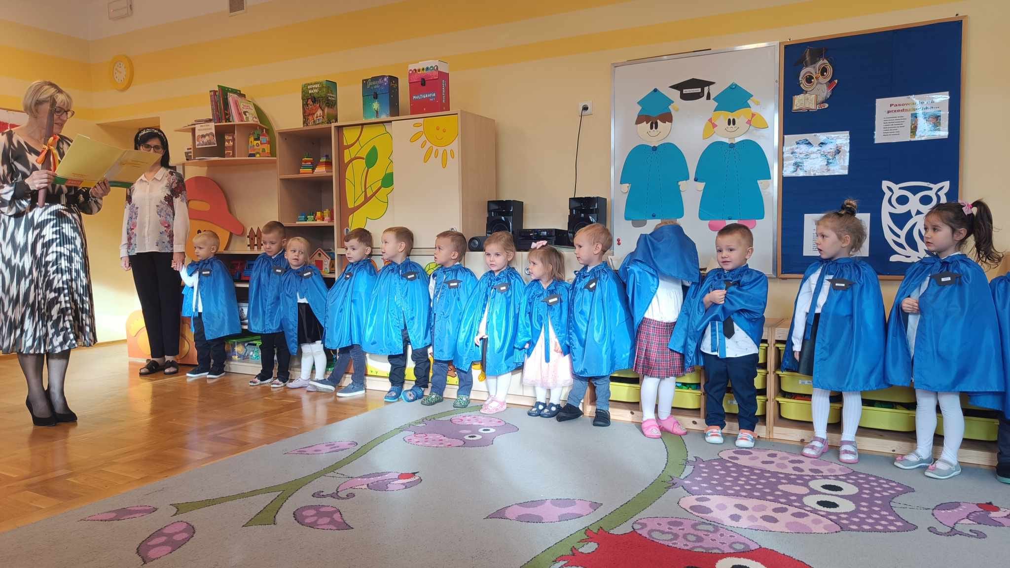 Dzieci stoją w p&oacute;łkolu w niebieskich pelerynkach.