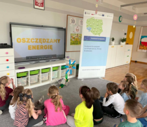 Dzieci siedzą na dywanie przed tablicą interaktywną.