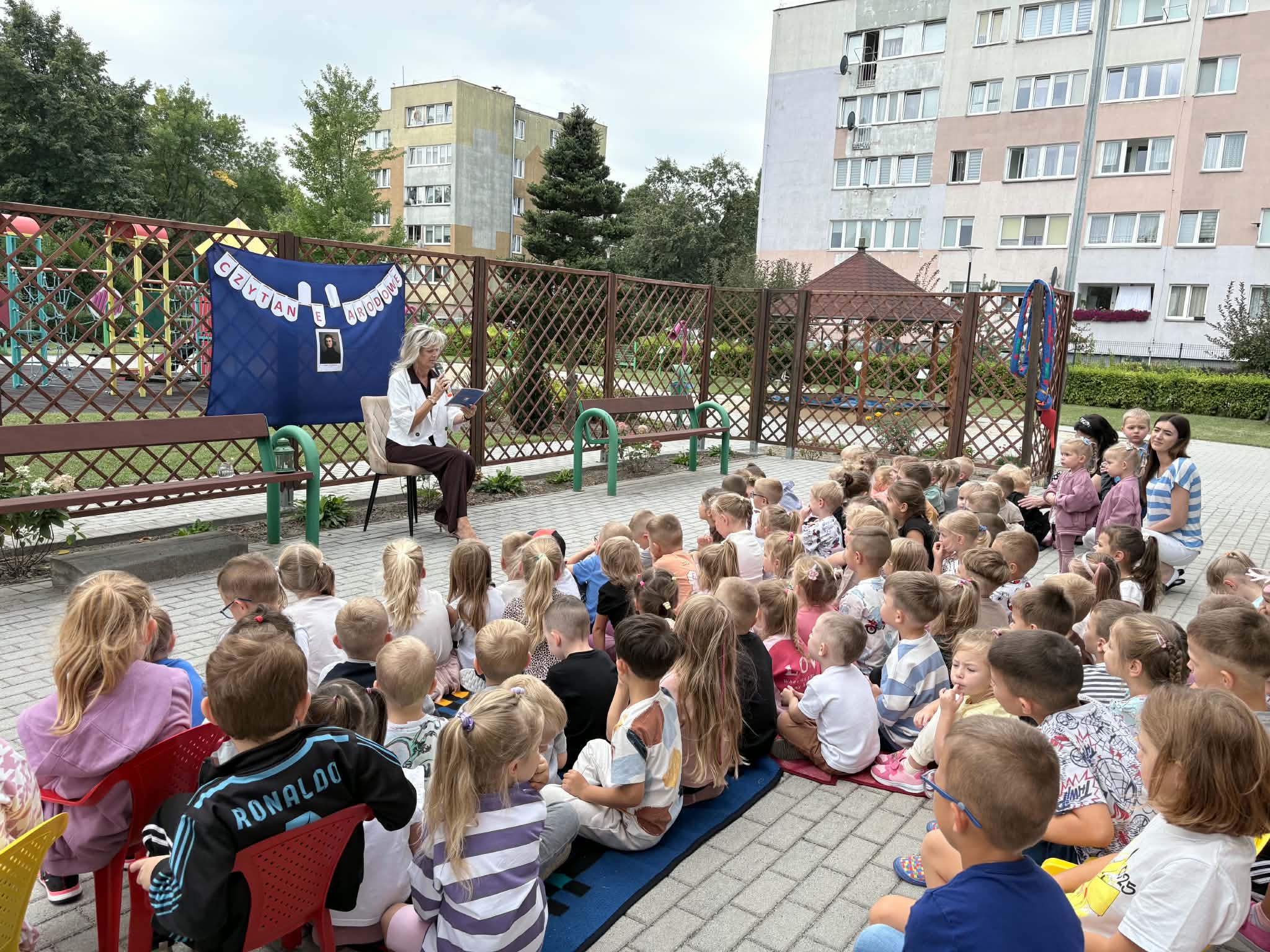 Dzieci siedzą na kocach na tarasie, nauczyciel czyta fragmenty książki.