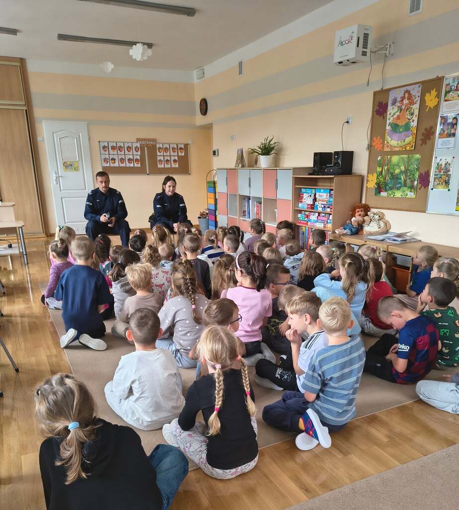 Dzieci siedzą na dywanie w grupie i słuchają policjant&oacute;w.