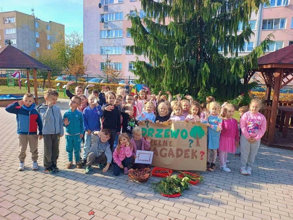 Dzieci stoja w grupie na dworze trzymając w ręku plakat z napisem drzewo pełne zagadek.