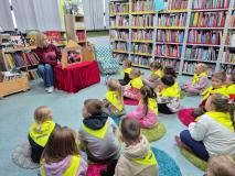 Dzieci siedzą na małych dywanikach w bibliotece i słuchają pani bibliotekarki.