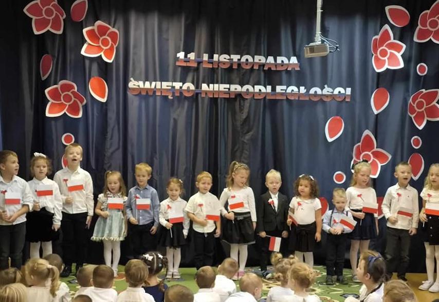 Dzieci ubrane na galowo trzymają w ręku flagę Polski i śpiewają.