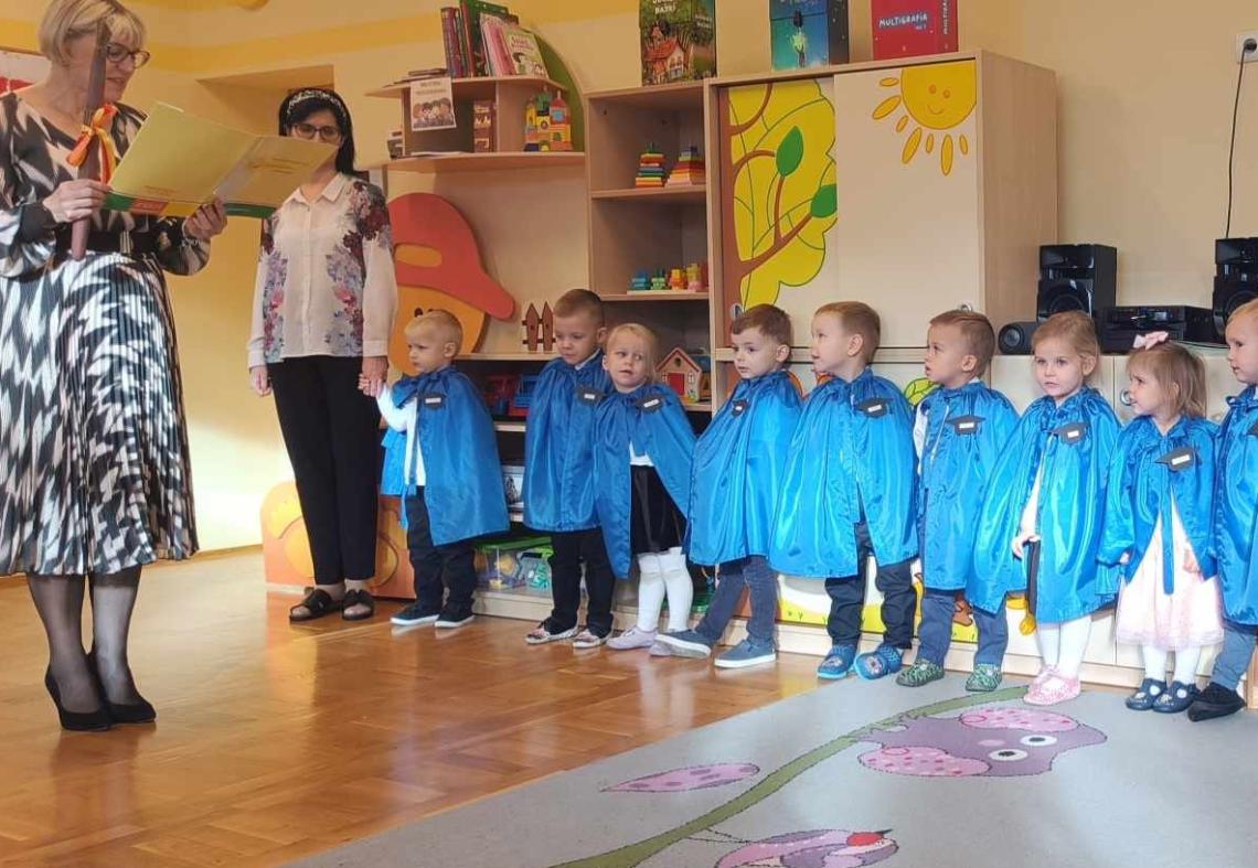 Dzieci stoją w półkolu w niebieskich pelerynkach.