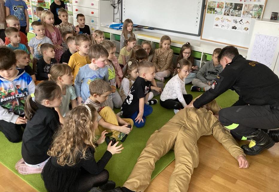 Dzieci siędza na dywanie dwóch strażaków pokazuje jak prawidłowo udzielać pierwszej pomocy.