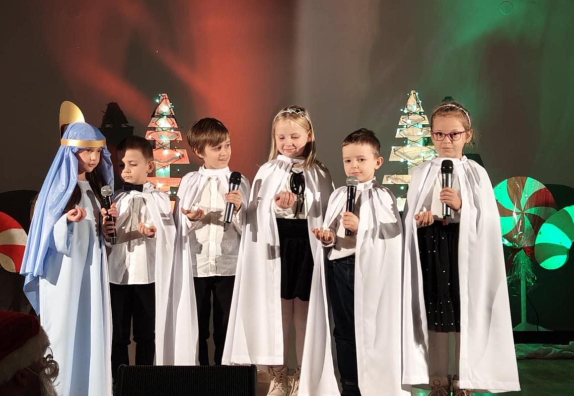 Pięcioro dzieci w białych pelerynkach stojąc na scenie śpiewają pastorłaki.