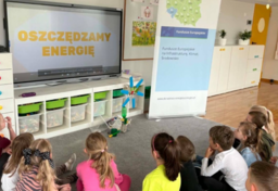 Dzieci siedzą na dywanie przed tablicą interaktywną.