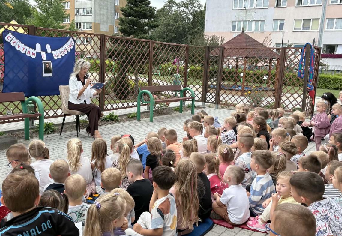 Dzieci siedzą na kocach na tarasie, nauczyciel czyta fragmenty książki.