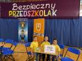 Dzieci ubrane w żółte koszulki siedzą przy stoliku na tle napisu bezpieczny przedszkolak.